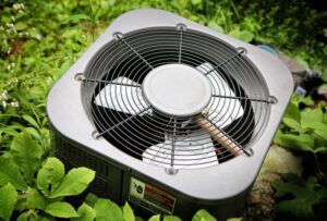 White outdoor HVAC condenser unit with metal fan grille surrounded by green landscaping and plants.