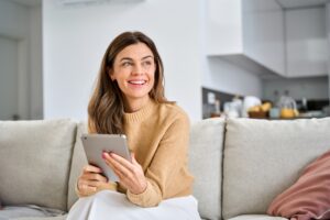 Woman in beige sweater sits on living room couch holding tablet with satisfied smile, modern kitchen visible in background.